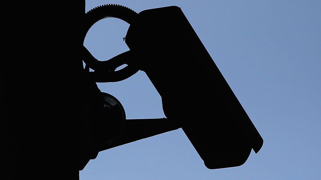 A silhouette of a CCTV camera outlined against a blue sky