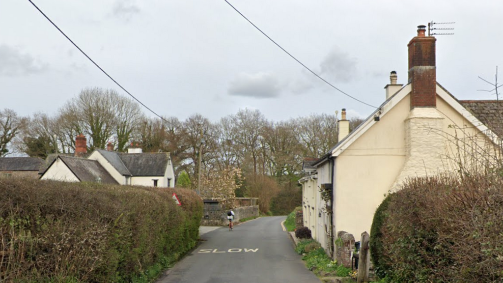 Google image shows Summer Lane near Teigngrace. Residential properties are built either side of the narrow road. A warning which states SLOW is painted on the road. A cyclist is travelling down the road.