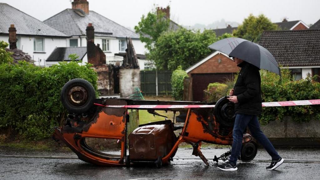 A man holding an umbrella walks past an overturned, burnt out car in Northern Ireland.