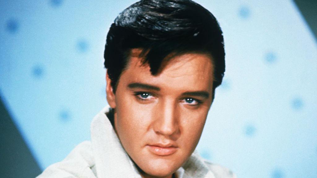 A portrait of Elvis Presley. He looks slightly away from the camera. He has dark hair and wears a white top with a large collar.