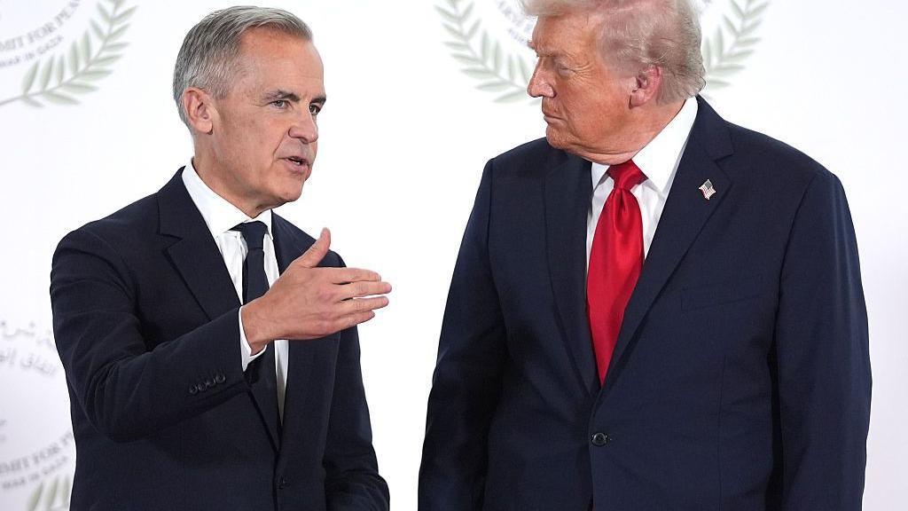 President Donald Trump greets Canada's Prime Minister Mark Carney during a world leaders' summit on ending the Gaza war on October 13, 2025 in Sharm El-Sheikh, Egypt. Prime Minister Mark Carney is on the left, wearing a black tie and a black suit. President Trump is on the right, wearing a dark blue suit and a red tie. PM Carney is gesturing as he speaks to Trump, who appears to be listening. Behind them is a white backdrop. 