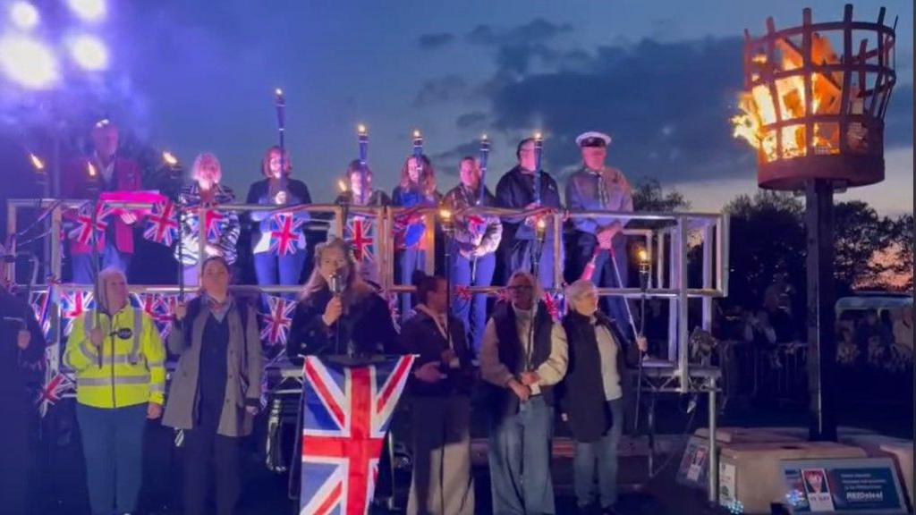 Two rows of people holding torches and standing on a platform decorated with union flags. In front of them a woman with a microphone is singing. Her music stand is decorated with a large union flag.