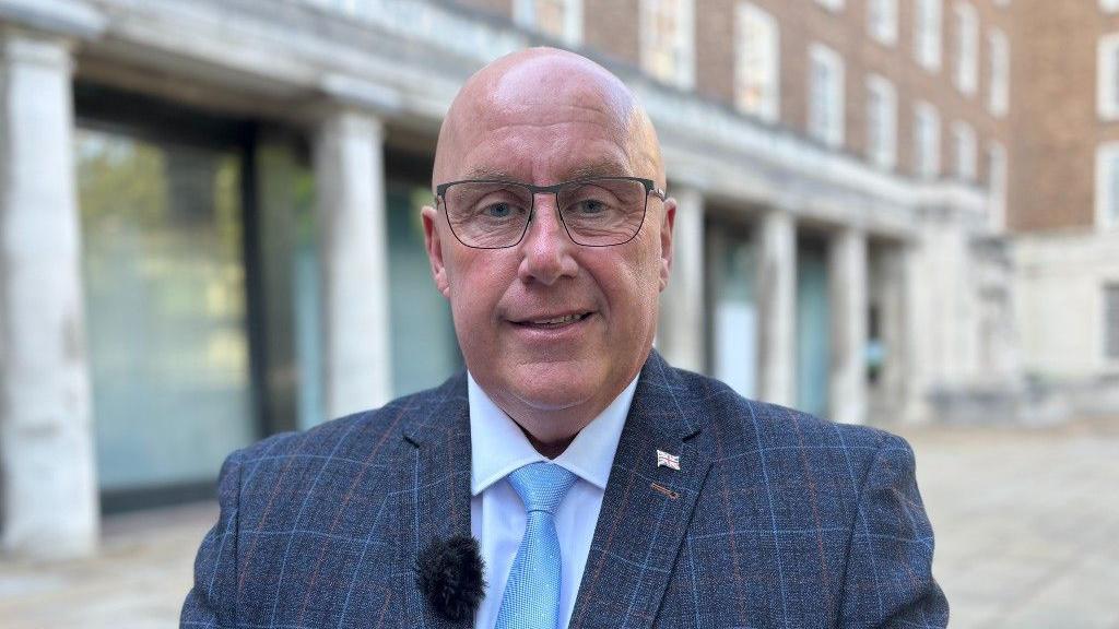 Mick Barton, Reform county council leader of Nottinghamshire, standing outside of County Hall wearing a blue, wool suit and light blue tie with a white shirt. He is a bald, middle-aged man wearing thin, metal-frames glasses.