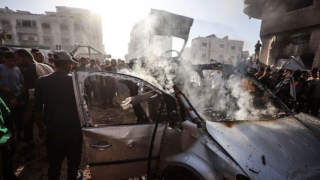 Crowds gather around a burnt out and destroyed car targeted by an Israeli airstrike in Gaza City. 