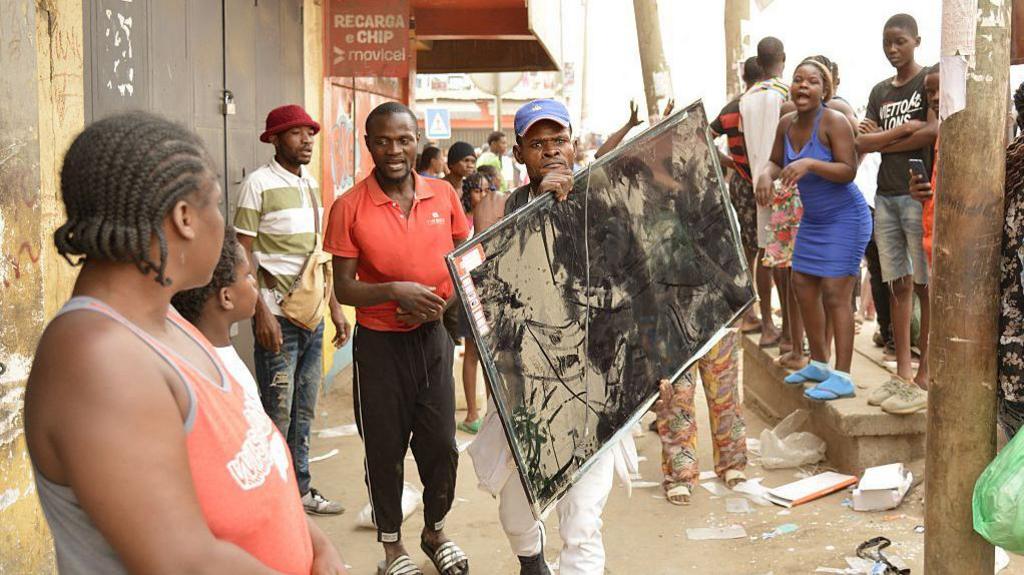 A man in a baseball cap carries a large-screen television as he walks down a street in Luanda. There are people lining the pavement looking at him. 
