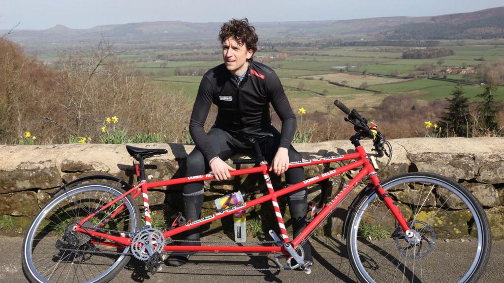 Greg sits on a stone wall with a red tandem bike leaning against him.