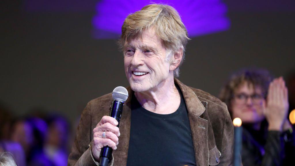 Robert Redford photographed at the age of 84. He is speaking into a microphone.