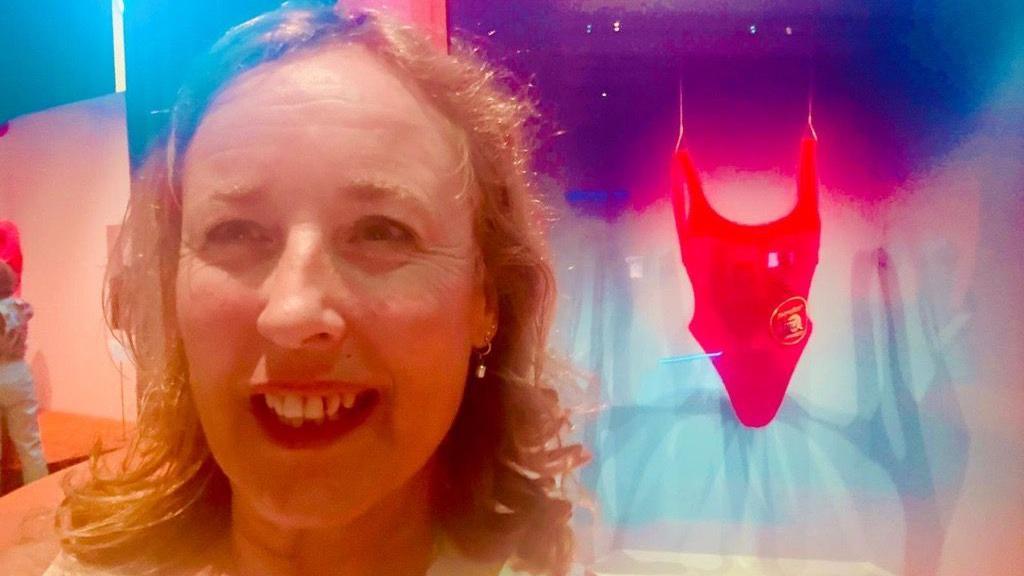 Shan taking a selfie in front of a red swimsuit  believed to be worn by Pamela Anderson inside a museum.
