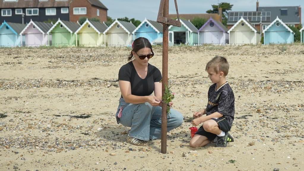 Harriet Whiteside, wearing a black top and blue denim jeans, kneels down tying some wild flowers to a rusty metal sign post. Beside her is a young boy kneeling wearing a dark-coloured patterned football kit. They are on the beach, with beach huts and houses in the background.
