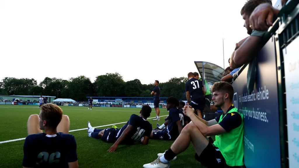 Des Buckingham, stood in a blue T-shirt and blue shorts, watches on as Oxford United play Oxford City at Court Place Farm in Marston. Oxford United players, wearing blue T-shirts, are sat around or lying at the side of the pitch.