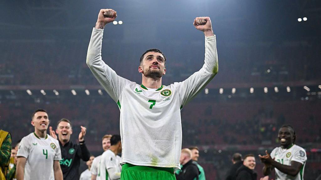 Troy Parrott of Republic of Ireland celebrates after the FIFA World Cup 2026 Group F Qualifier match between Hungary and Republic of Ireland at Puskás Aréna in Budapest, Hungary. He has short dark hair wearing his white and green Ireland kit. He is holding up both fists in the air. Other footballers are in the background smiling at him and clapping.