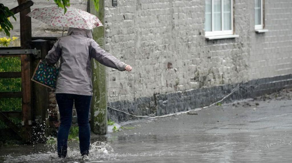 A pedestrian holding under an umbrella while walking away from the camera negotiates a partially flooded footpath