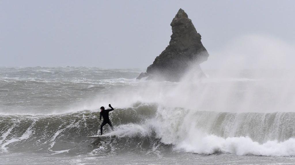 A surf going down the line on an overhead wave. The wave is breaking with white foam which the wind is spraying high into the air. In the back round there is a steep rock jutting out of the water 