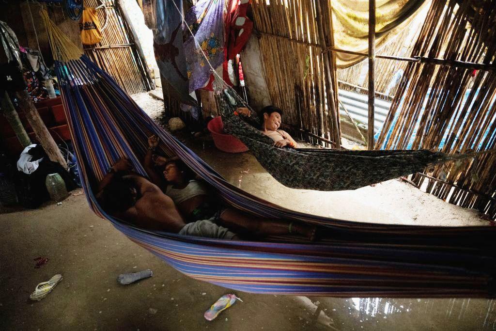 Two people lying in one hammock, and another in a separate hammock, inside a room built from wooden poles. There is shallow water on the ground below the hammocks, with two sandals floating in the water.