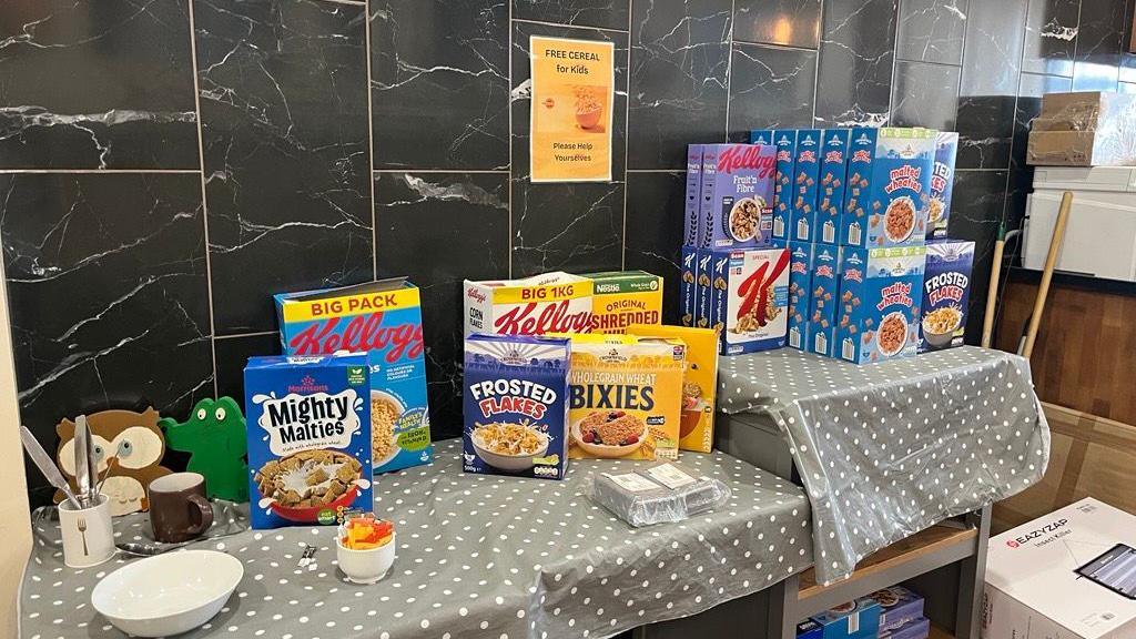 A table with a number of boxes of cereal on, as well as a grey polka dot tablecloth. A sign on the wall reads "Free cereal for kids. Please help yourselves".