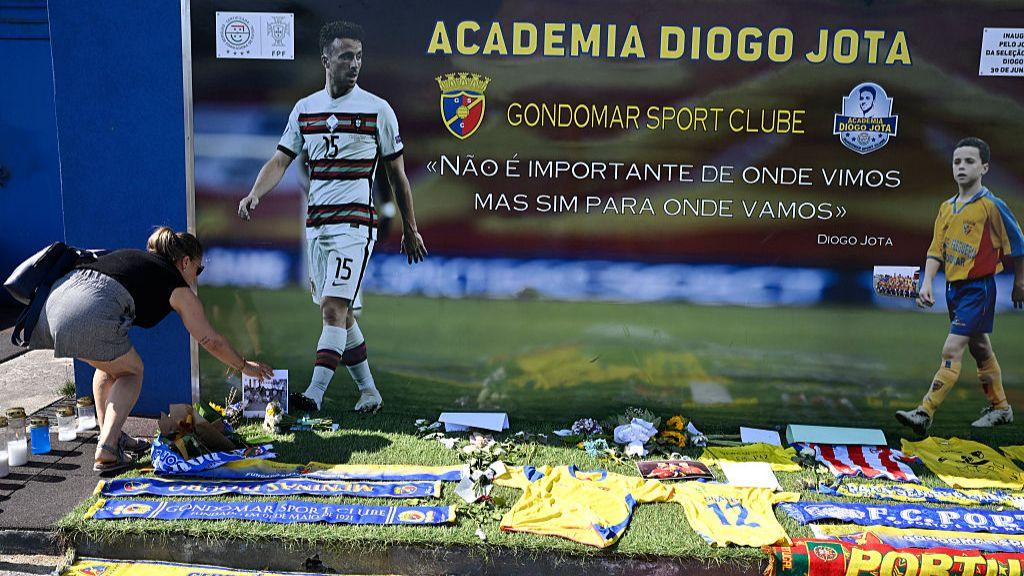 A woman in a short jeans skirt and black tank top lays flowers outside the Academy Diogo Jota in Gondomar. A poster of the player as an adult and as a child in his local team kit hangs off the external walls, shirts and football scarves lay on the ground in front of it