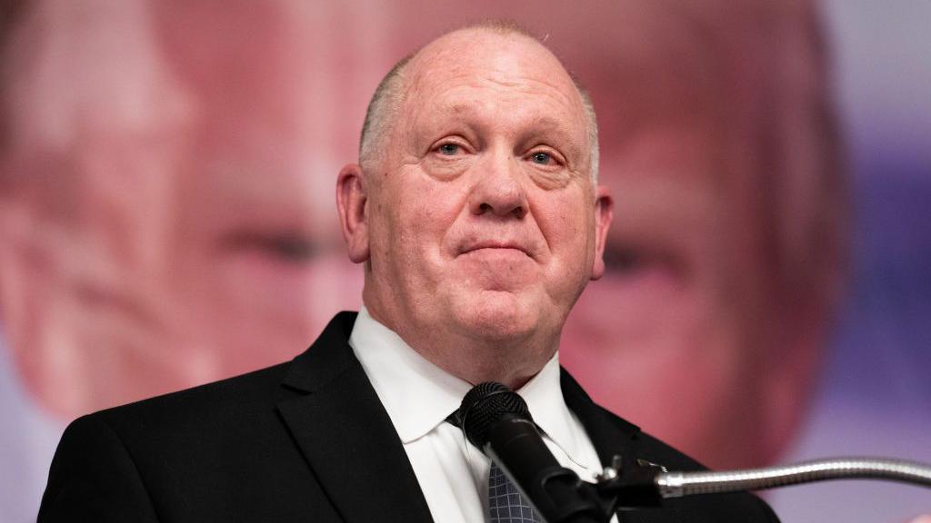 Tom Homan, a man with short white/grey hair, wears a black jacket and white shirt, standing in front of a microphone with an image of Donald Trump behind him 