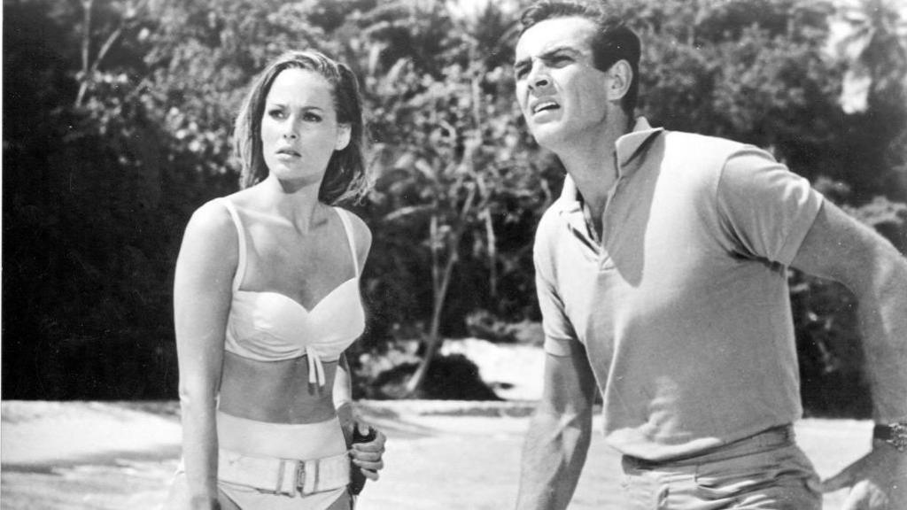 A black and white still image from the film Dr No. A young Sean Connery and a young Ursula Andress are looking off camera. They are on a beach with trees behind them. She is wearing a white bikini and he is in trousers and a T-shirt
