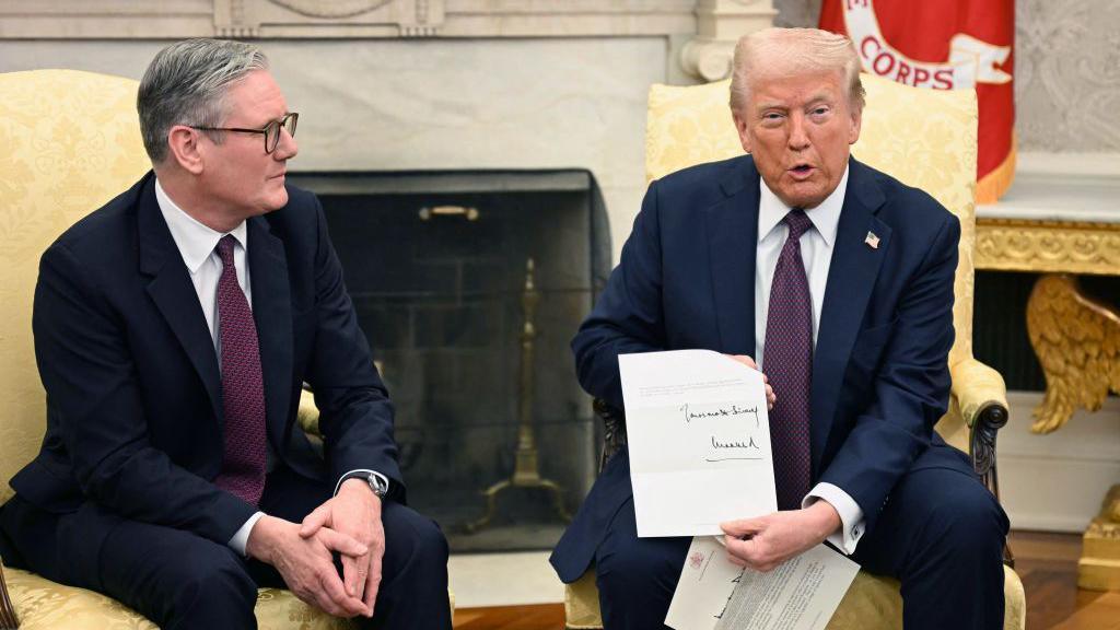 US President Donald Trump holds a letter from Britain's King Charles III during a bilateral meeting with British Prime Minister Keir Starmer in the Oval Office. Both men wear dark blue sits and sit on cream-upholstered wing chairs