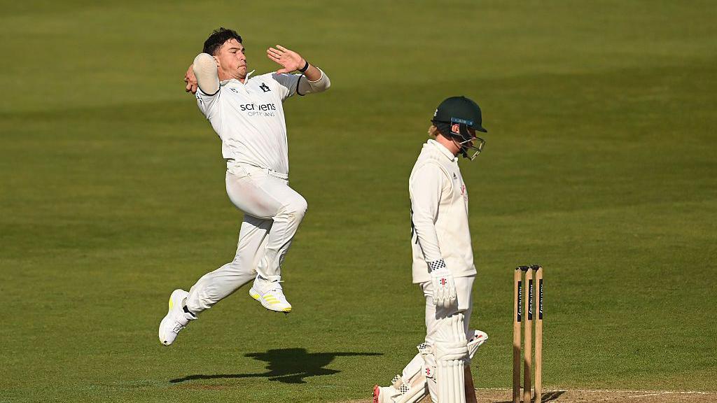 Michael Booth leaps off the ground in take-off as he prepares to bowl. Notts batsman Kyle Verreynne is seen preparing to back-up.