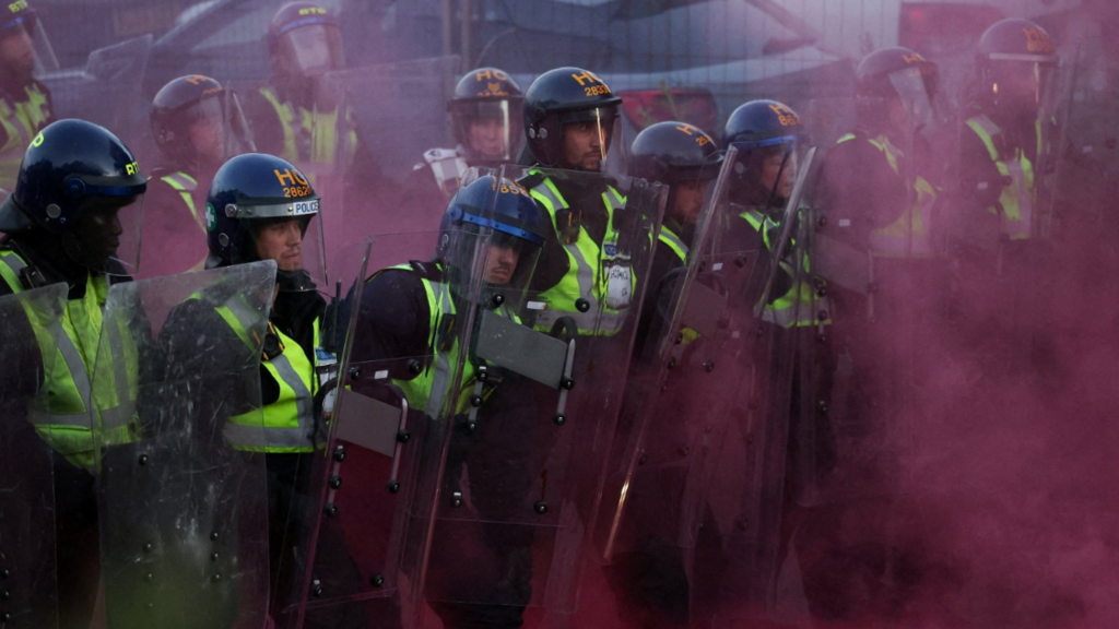 A line of police officers wearing riot gear are surrounded by red smoke, probably from a flare