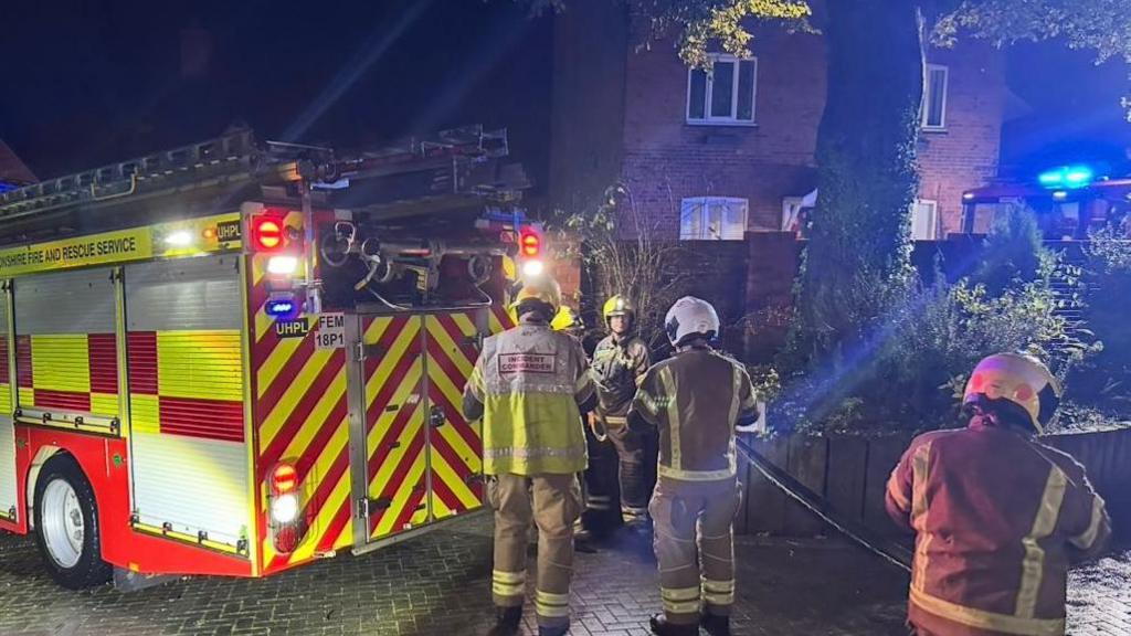 Four firefighters wearing protective uniforms and helmets are standing next to a fire engine - two of them are holding a hose. Behind them is a two-storey brick-built house.