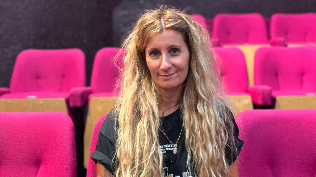 Woman with long blonde hair and a black t-shirt on pink chairs smiling.
