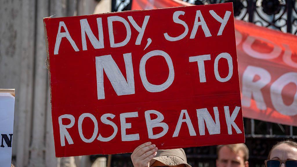 A person holding a sign that is red and in white writing it says "Andy, say no to Rosebank".