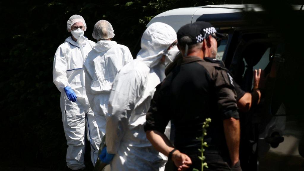 Forensic team members in white protective suits stand with police officers outside a home