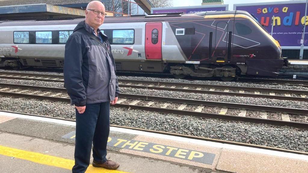 Railfuture spokesperson Dave Richardson at Oxford Train Station. There is a CrossCountry train on the track behind him. It's a cloudy day.