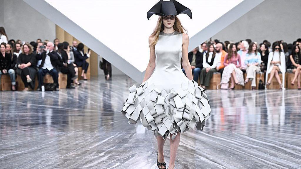 A woman in a silver dress with a black hat. She walks on a catwalk. The audience are visible behind her.
