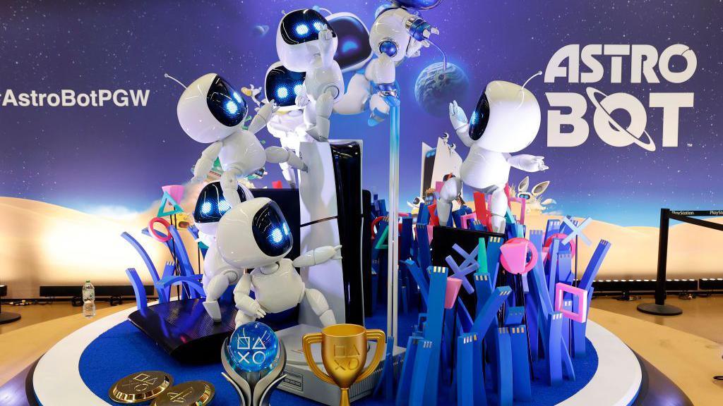 A large model of several curvy white robots standing around a PS5 console, with some jumping up in the air. There are trophies surrounding them.