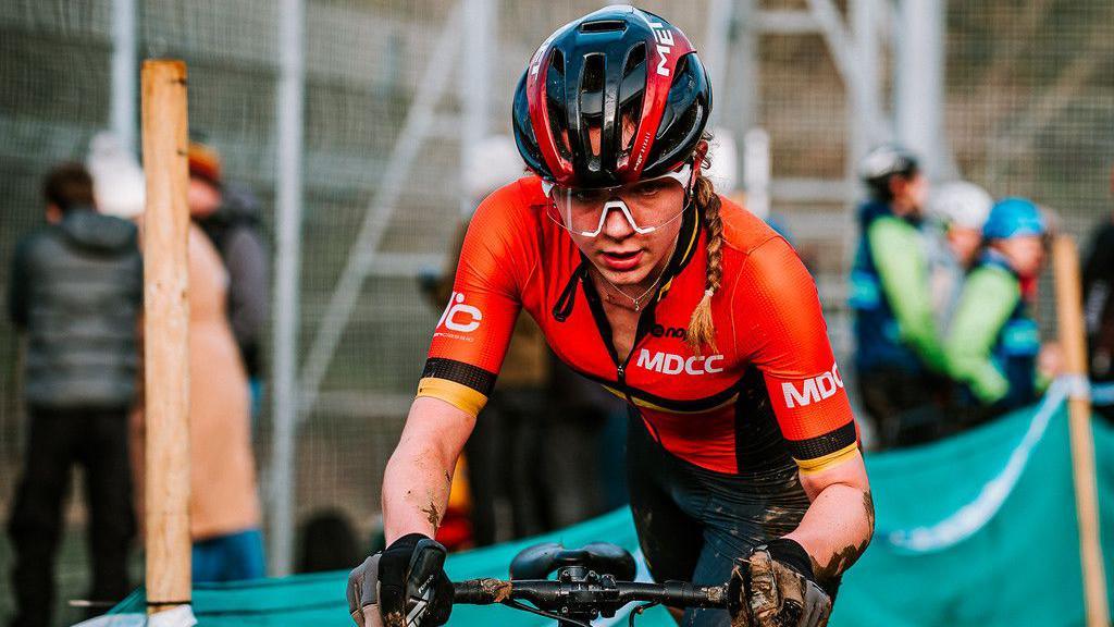 Lauren Fox, wearing a helmet, orange cycling top and black cycling shorts, looks determined as she guides her bike through a course. 