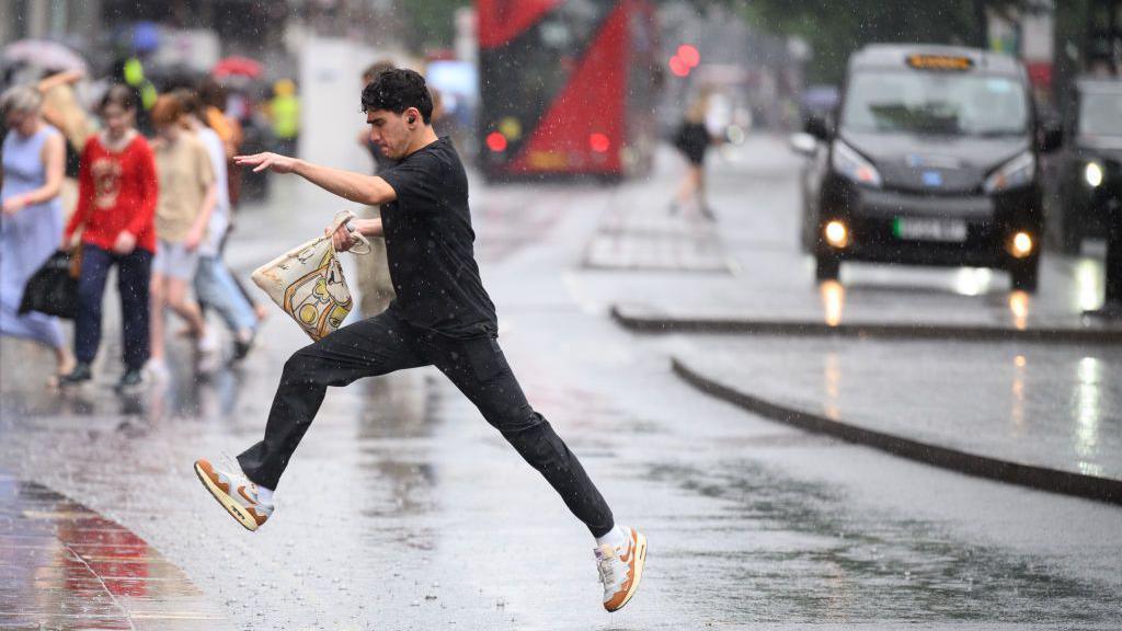 A young man jumps over a large puddle in the road. It is raining. Other pedestrians, a London bus and a black taxi are in the background.