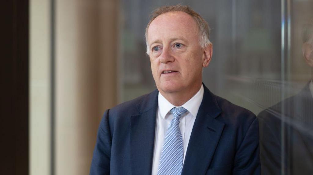 John Neal, former chief executive officer of Lloyd's of London, leaning against a glass wall in an atrium outdoors. He is wearing a navy blue suit, white shirt, and patterned white and blue tie.