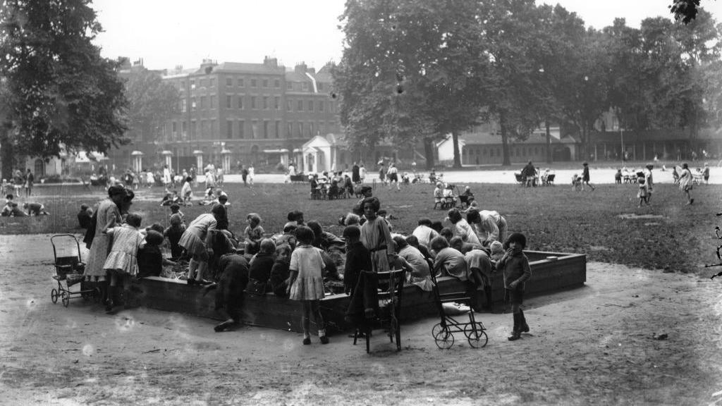 A black and white image shows children playing in a sandpit at the Foundling Hospital in 1930, with girls lin light-coloured short dresses surrounded by metal dollies' prams, and trees and the hospital in the background