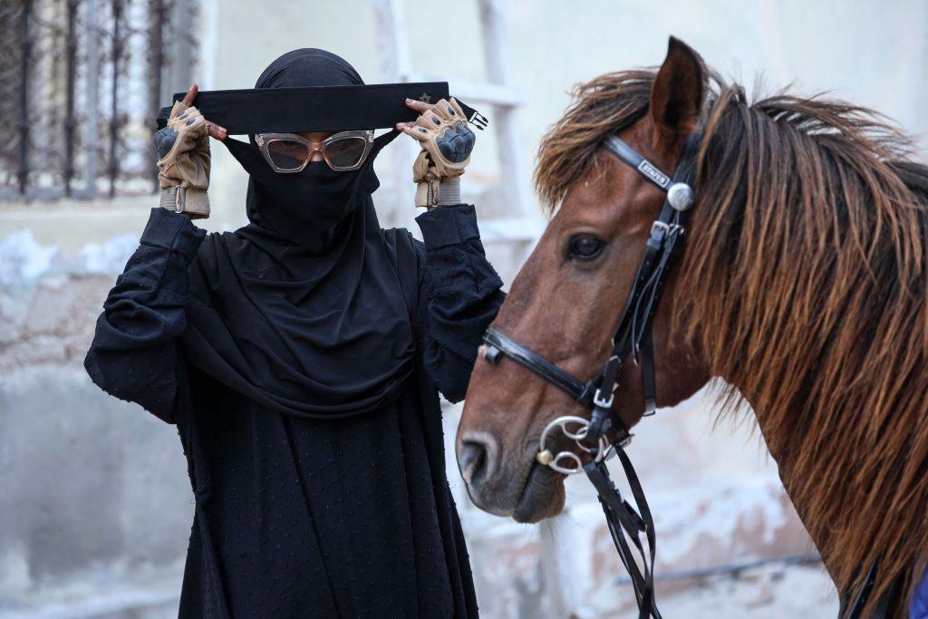 Woman wearing sunglasses and Islamic dress - including an abaya and niqab - gets ready to mount her chestnut-coloured horse.