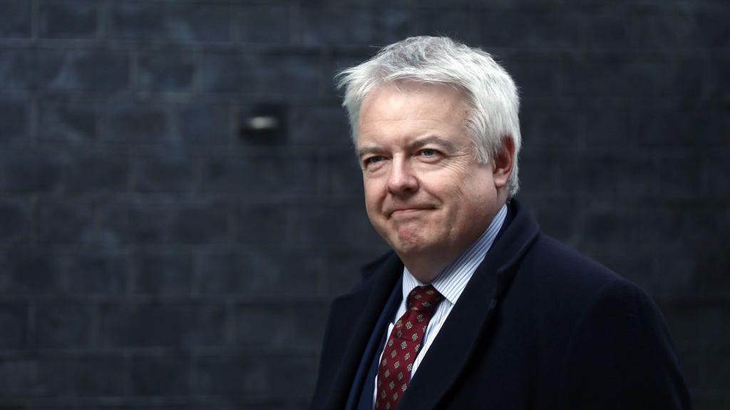 Carwyn Jones, who has grey hair, wears red patterned tie, vertically striped shirt and a dark coat. Behind him is a grey brick wall