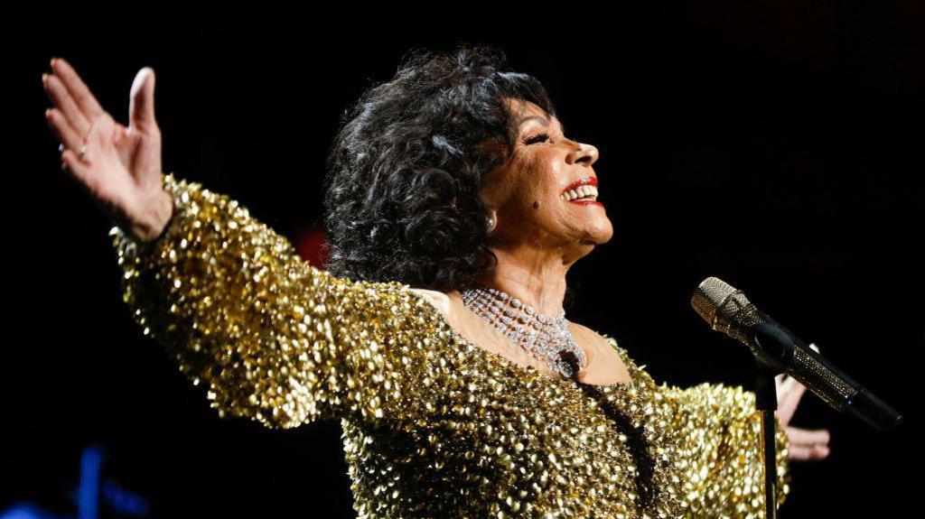 Dame Shirley Bassey performs on stage accompanied by The Royal Philharmonic Concert Orchestra during The Sound of 007 in concert at The Royal Albert Hall on October 04, 2022. She is wearing a gold sparkly dress and her arms are outstretched, she is smiling at the crowd. 