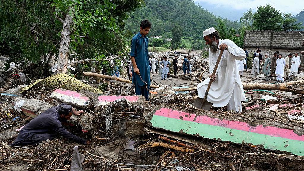 A man in white robes and hat shovels among debris, alongside a young boy in blue robes and another man in darker robes down on all fours and digging in the mud