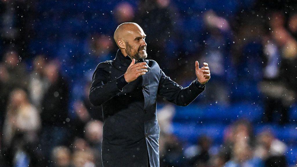 Luke Williams smiling and looking to the right with both his arms raised. He is wearing a long, dark coat. There is a mist of rain in the air and fans sitting in the stands in the background, which is blurred.