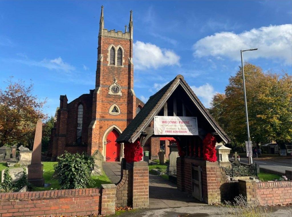 A church with orange bricks and a red door, surrounded by graves and a brick wall. There are trees to the right and one on the left.