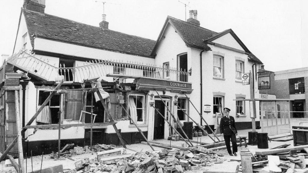 A police officer stands in front of the debris at the Horse and Groom with the ground floor front windows blown in