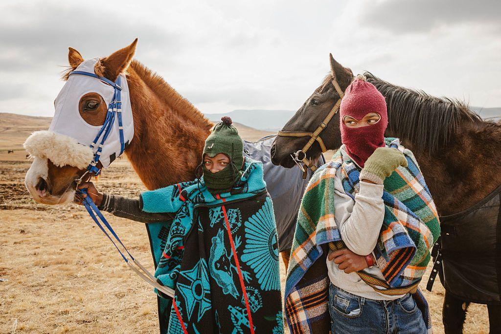 Two jockeys stand next to their horses. They both wear balaklava and are wrapped up in blankets.