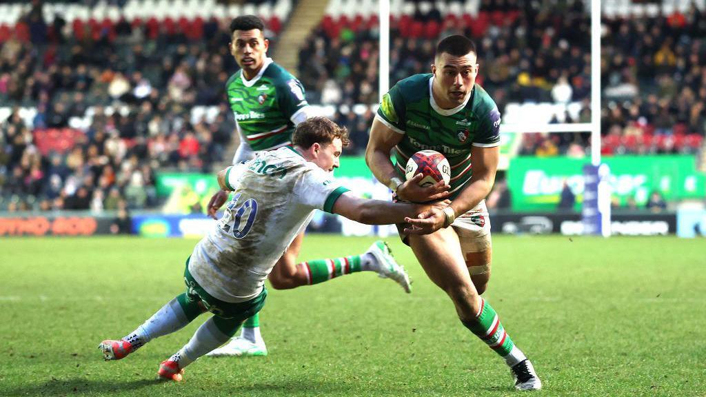 rugby player in green holding rugby ball darts past tackling player in white 