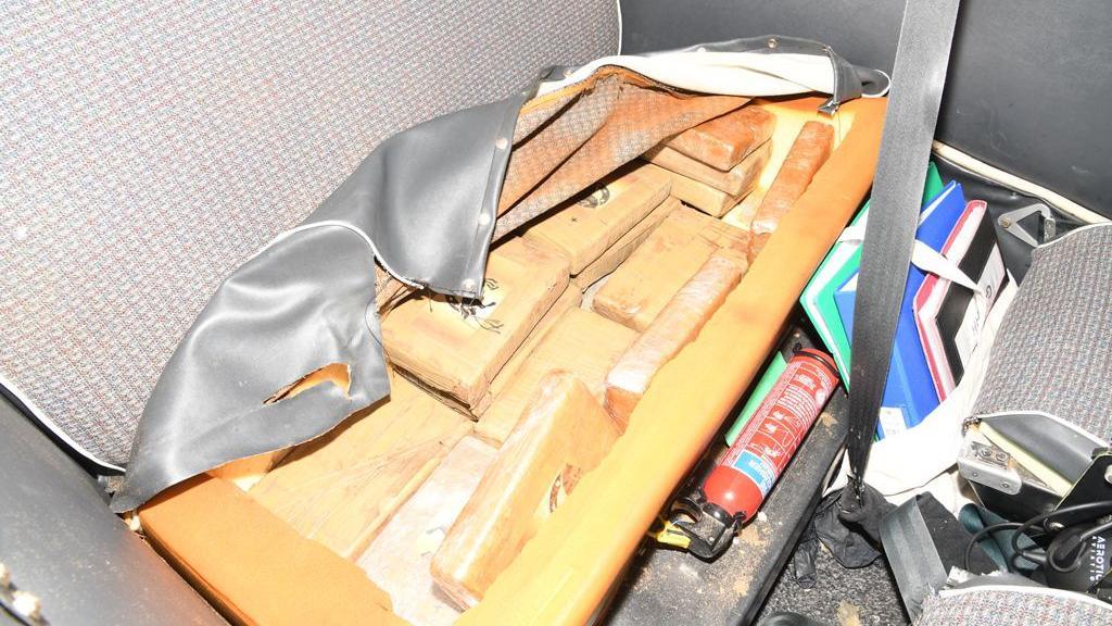 Brown packages are hidden in a grey seat. On the floor are multiple notebooks and a seat belt. A small red fire extinguisher is also on the floor. 