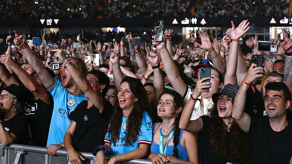 Oasis fans at the Principality Stadium. A large number of people can be seen looking towards the stage. Many have their hands in the air and are recording the concert on their smartphones.