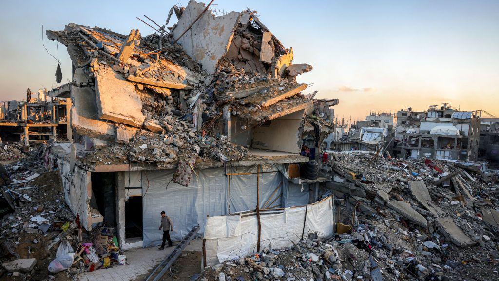 The remnants of a collapsed building can be seen with a man walking in front, with the rest of the buildings around them almost all completely flattened