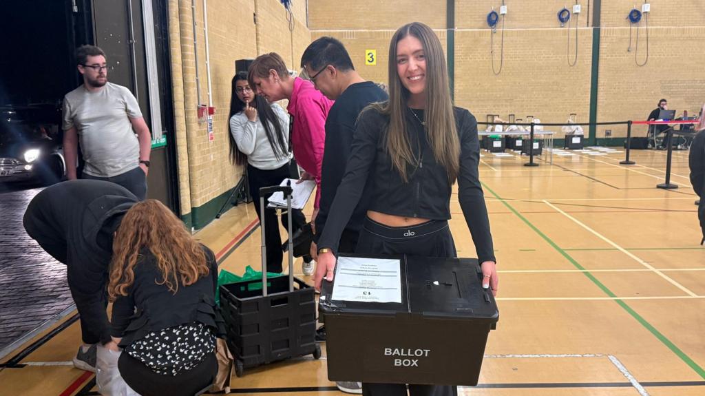 A woman holding a ballot box in a sports hall.