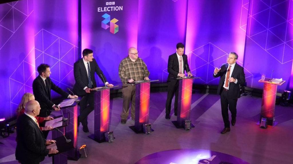 Six politicians standing in a line behind lecterns in a TV studio